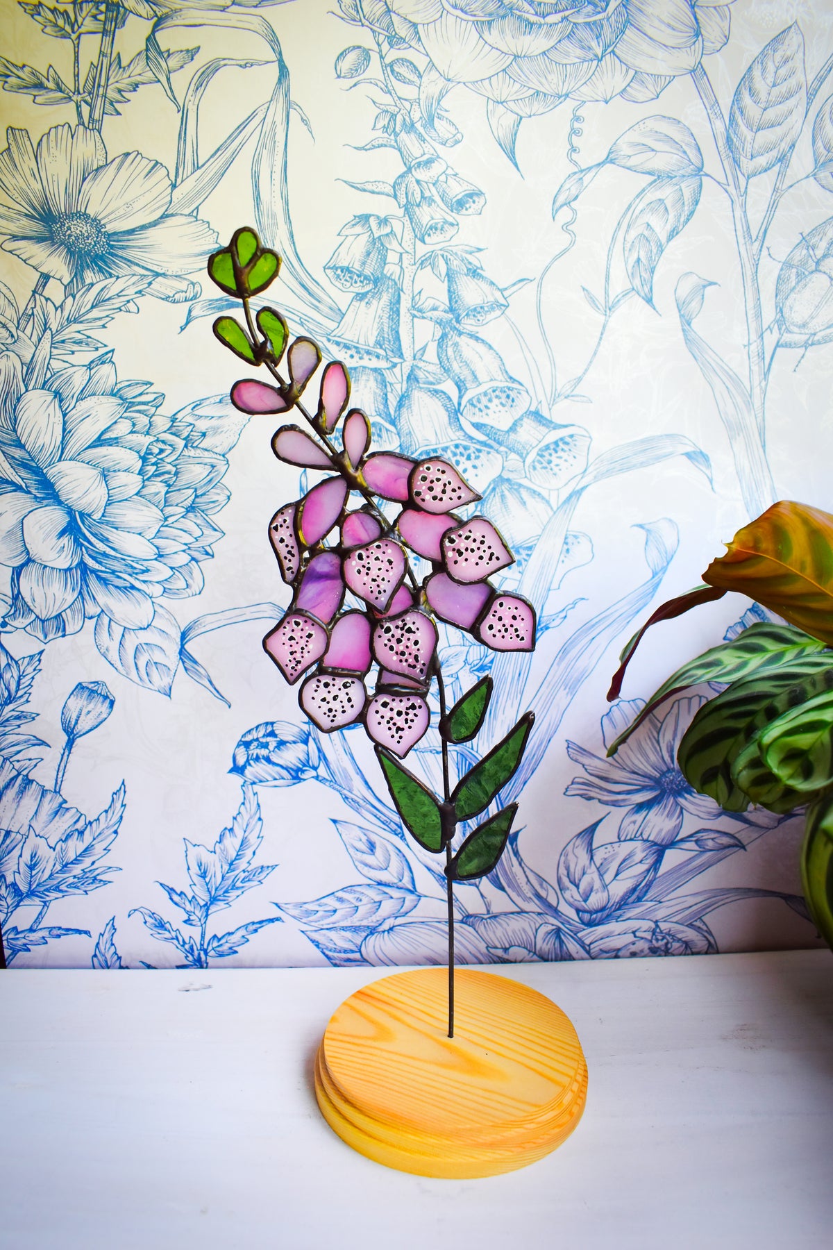 Pink stained glass foxglove flowers with green leaves on a wooden stand beside a window.”