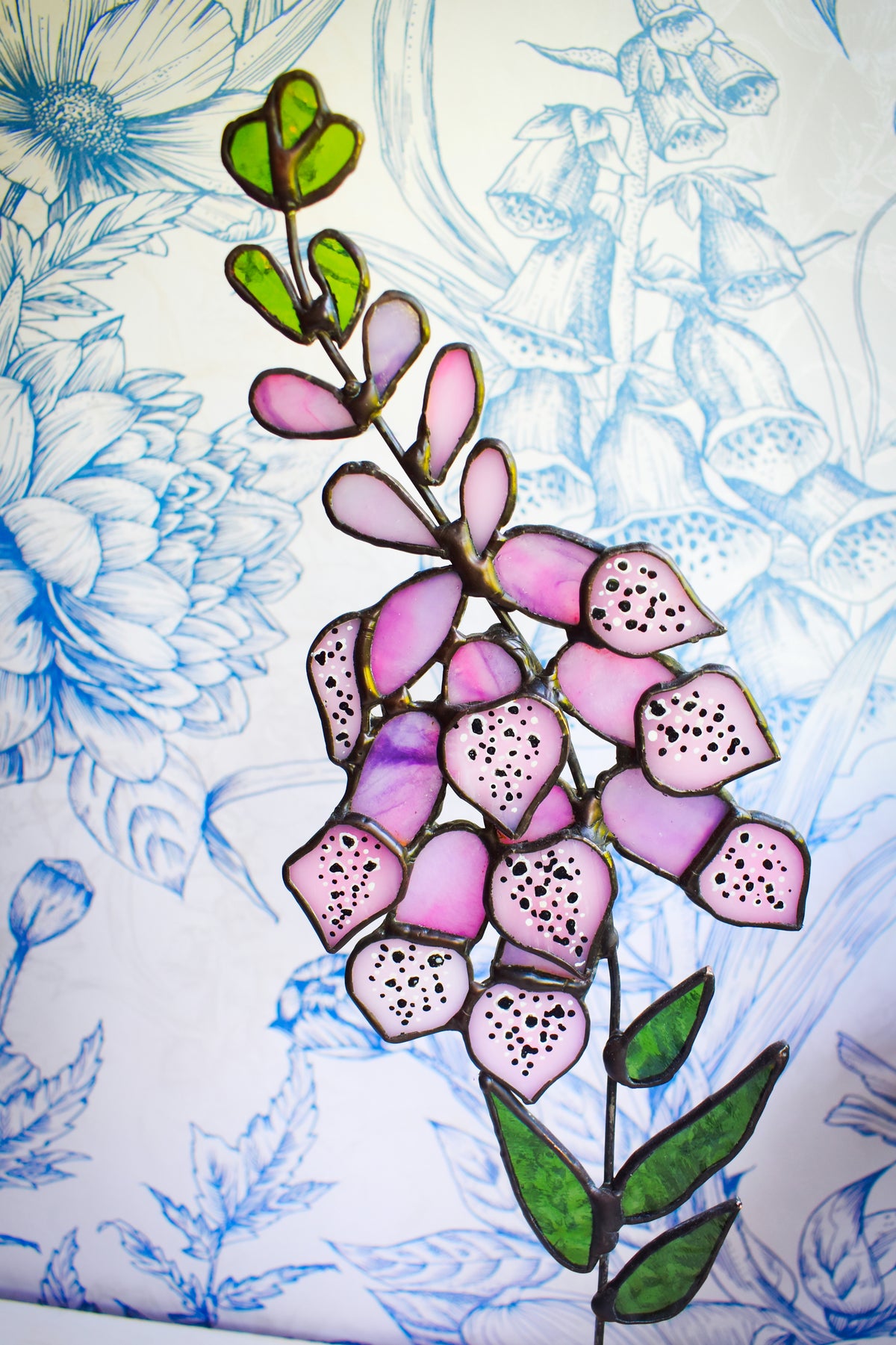 Pink stained glass foxglove flowers with green leaves on a wooden stand beside a window.”